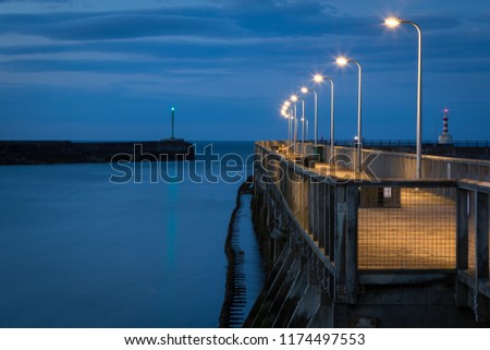 Wooden pier in the fishing village Amble, Northumberland, UK. Evening scene,  footpath illuminated by row of lamp posts. Royalty-Free Stock Photo #1174497553