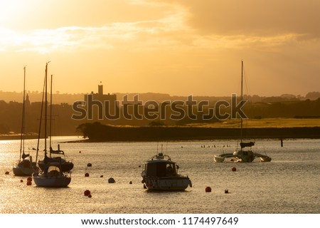 Beautiful summer sunset landscape in Amble town, Northumberland, UK. Yellow and orange sky above water and marina, Warkworth Castle in background. Royalty-Free Stock Photo #1174497649