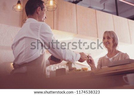 Smile and pay. Joyful pretty aged woman standing opposite a man smiling and holding a credit card.