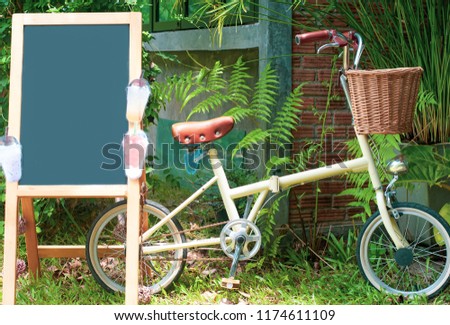 An empty blackboard on wooden stand ready for put text in, besides of its is beautiful foldable bike, background is green plant