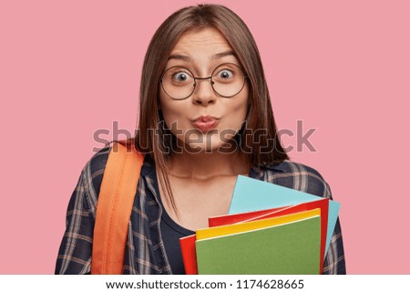 Shot of beautiful young female reader keeps lips pressed, has lovely gaze at you, flirts with boyfriend, holds pile of scientific books, carries orange rucksack on back, isolated over pink background