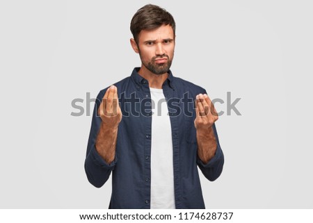 Handsome brunette man with thick dark beard makes money gesture, thinks about profitable business, demonstrartes symbol of currency and richness, tries to specify his mind. Body language concept