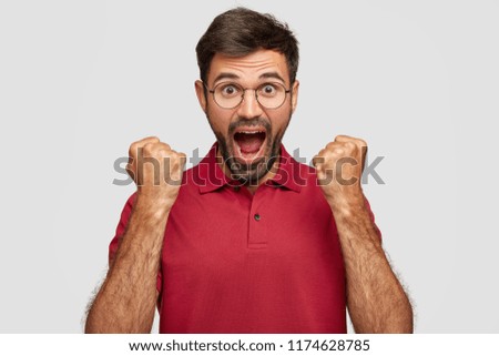 Emotional bearded guy keeps hands clenched in fists, exclaims loudly, has nervous expression as shouts for favourite football team, dressed in casual clothes, isolated over white background.