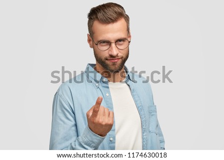 Body language concept. Attractive young man with stylish haircut, looks with appeal at camera, says come here, gestures with index finger, has mysterious expression, isolated on white background Royalty-Free Stock Photo #1174630018