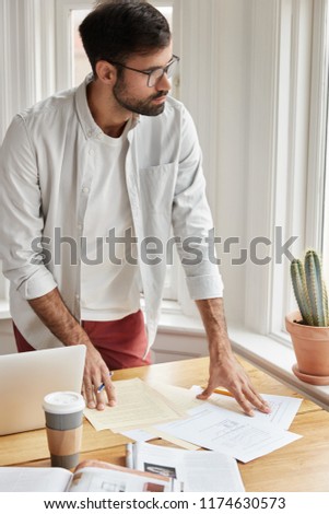 Vertical shot of male coworker cant make right accountings in papers, looks at documents planner, stands at desktop, wears round glasses for good vision, drinks fresh beverage, uses free internet