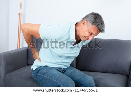 Mature Man Sitting On Sofa Suffering From Back Pain Royalty-Free Stock Photo #1174636561