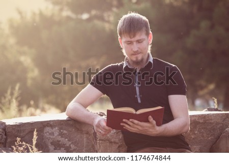A man reading a book sitting on a bench in a park.