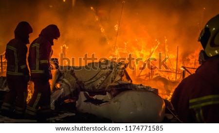 Firemen in professional uniform in front of the burning structure, fire of a falla sculpture made of paper at Las Fallas festival in Cullera, Valencia, Spain.