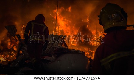 Firemen in professional uniform in front of the burning structure, fire of a falla sculpture made of paper at Las Fallas festival in Cullera, Valencia, Spain.