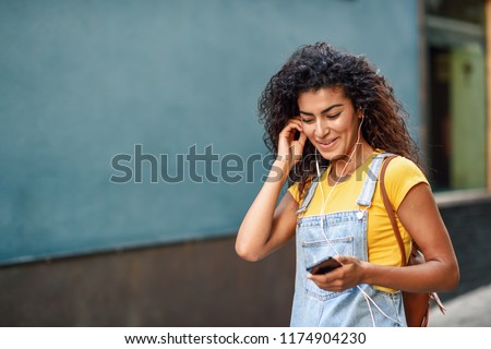 Attractive African woman listening to music with earphones outdoors. Black girl in casual clothes with curly hairstyle in urban background.