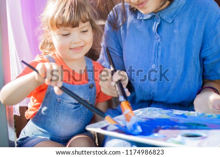 Young artist teacher drawing a picture with her little student , holding brushes and palette with blue paint. Art lessons for children, art school
