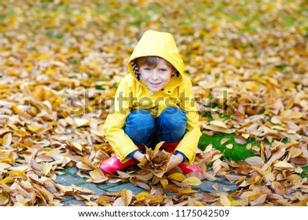 Portrait of happy cute little kid boy in yellow rain coat and red rubber boots with autumn leaves background. Funny child having fun and playing in fall forest or park on cold autumnal day