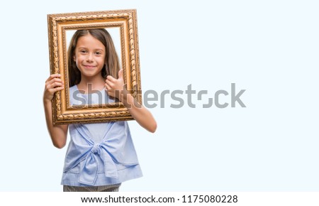 Brunette hispanic girl holding vintage art frame happy with big smile doing ok sign, thumb up with fingers, excellent sign