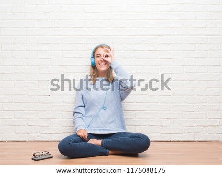Beautiful young woman sitting on the floor listening music wearing headphones at home with happy face smiling doing ok sign with hand on eye looking through fingers
