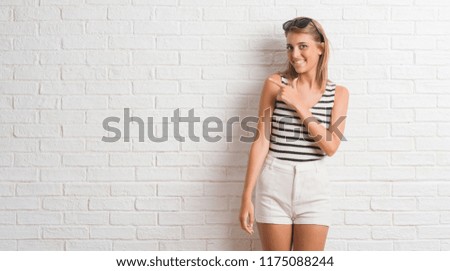 Young beautiful blonde woman over white brick wall cheerful with a smile of face pointing with hand and finger up to the side with happy and natural expression on face looking at the camera.