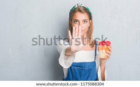 Beautiful young woman over grunge grey wall eating fruits with open hand doing stop sign with serious and confident expression, defense gesture