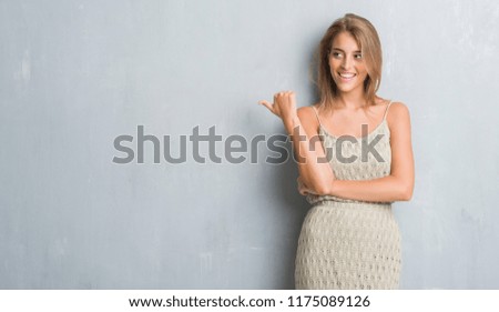 Beautiful young woman standing over grunge grey wall pointing and showing with thumb up to the side with happy face smiling