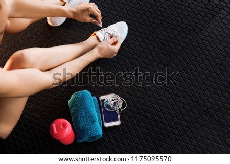 woman tying shoelaces of white trainers siting on a mat surrounded by mobile phone, earphones, towel and a shaker in the gym. top view Royalty-Free Stock Photo #1175095570