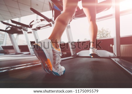 close up womans legs running at the treadmill in the gym against the sunrise. lady wearing white orange blue sneakers. Cardio exercise. concept of healthy lifestyle Royalty-Free Stock Photo #1175095579
