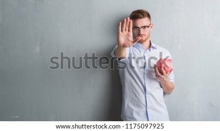 Young redhead man over grey grunge wall holding piggy bank with open hand doing stop sign with serious and confident expression, defense gesture