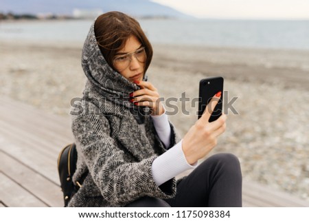 stylish, daring dark-haired girl in a gray coat and glasses takes pictures of the seascape