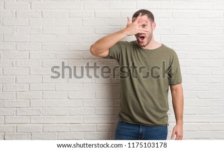 Young caucasian man standing over white brick wall peeking in shock covering face and eyes with hand, looking through fingers with embarrassed expression.