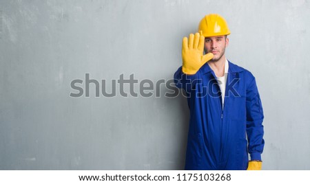 Young caucasian man over grey grunge wall wearing contractor uniform and safety helmet with open hand doing stop sign with serious and confident expression, defense gesture