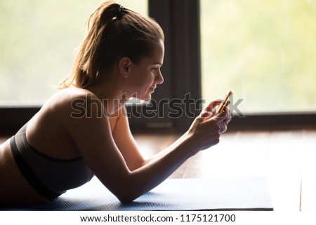 Young sporty smiling woman after practicing yoga, enjoy a break in doing exercise, relaxing on yoga mat, using sport app on her mobile phone, messaging, indoor close up, yoga studio