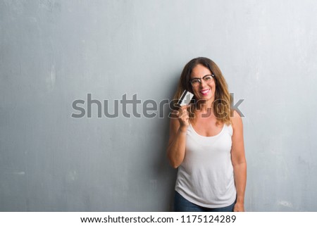 Middle age hispanic woman standing over grey grunge wall holding credit card with a happy face standing and smiling with a confident smile showing teeth
