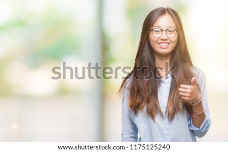 Young asian business woman wearing glasses over isolated background doing happy thumbs up gesture with hand. Approving expression looking at the camera with showing success.