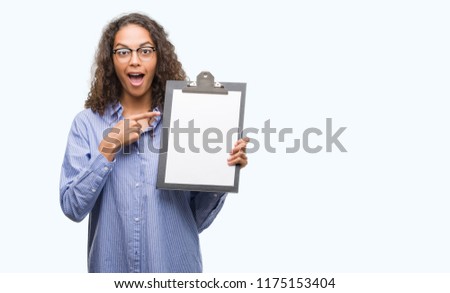 Young hispanic business woman holding clipboard very happy pointing with hand and finger