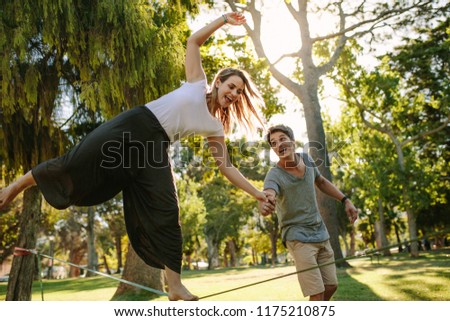 Woman doing slack rope walking in a park. Man helps woman in balancing during her tightrope walk in a park. Royalty-Free Stock Photo #1175210875