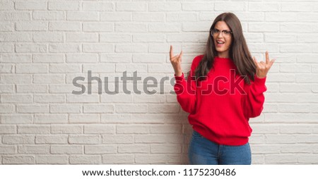 Young brunette woman standing over white brick wall shouting with crazy expression doing rock symbol with hands up. Music star. Heavy concept.