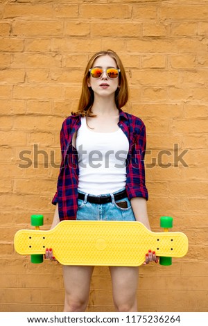 A girl wearing sunglasses, checkered shirt and denim shorts is standing in front of the brick wall and holding a yellow longboard in front of her. Cool style.