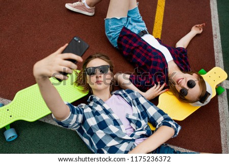 Two pretty blond girls wearing checkered shirts, caps and denim shorts are lying on the bright longboards on the sportsfield with their sunglasses on and making selfie. Sport and cool style.