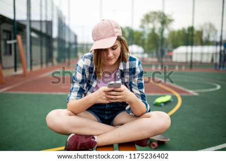 A pretty blond girl wearing checkered shirt, pink cap and denim shorts is sitting cross-legged on the sports field and looking at the phone. A green longboard is in the background.