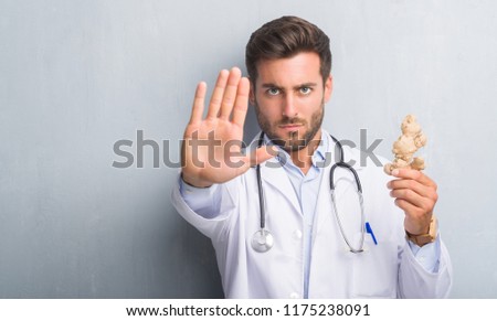 Handsome young doctor man over grey grunge wall holding ginger with open hand doing stop sign with serious and confident expression, defense gesture