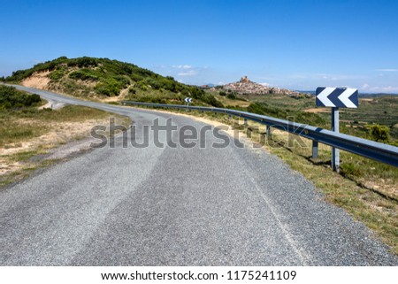 Spain, Navarre, Ujue: Panorama view with bending asphalt road, traffic sign, green hill and famous church of Santa María of old small Spanish village, green field and blue sky - concept travel