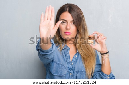 Young adult woman over grey grunge wall eating chocolate macarons with open hand doing stop sign with serious and confident expression, defense gesture