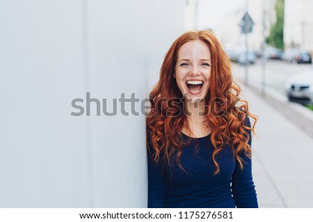 Cute young woman with a lovely sense of humour standing leaning against a white exterior wall with copy space in an urban street laughing at the camera Royalty-Free Stock Photo #1175276581