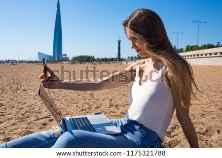 Woman university student photographing herself on cellphone during online learning on laptop computer while relaxing on the beach in sunny summer day. Hipster girl blogger having video conference 
