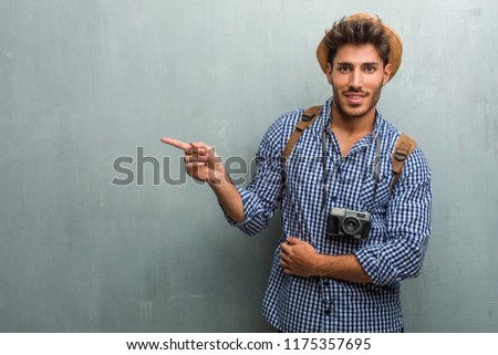 Young handsome traveler man wearing a straw hat, a backpack and a photo camera pointing to the side, smiling surprised presenting something, natural and casual