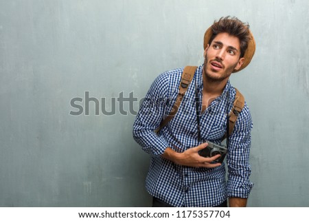 Young handsome traveler man wearing a straw hat, a backpack and a photo camera looking up, thinking of something fun and having an idea, concept of imagination, happy and excited