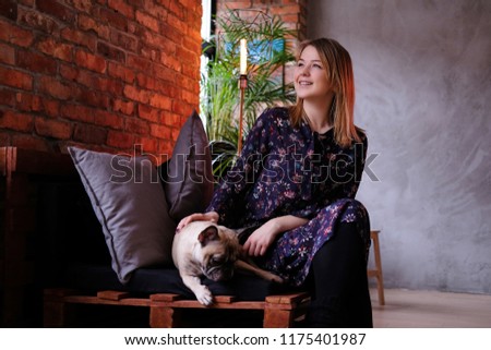 Happy blonde woman in dress sitting with her cute pug on a handmade sofa in room with loft interior.