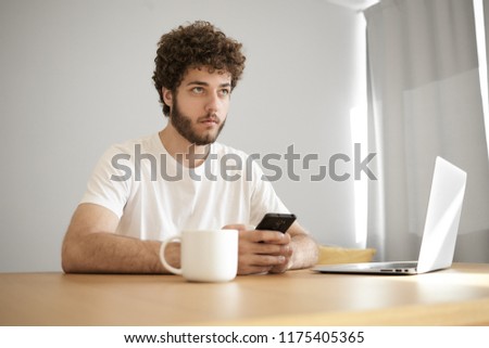 Always in touch. Picture of handsome stylish young European male blogger using modern electronic gadgets at wooden desk, drinking tea, checking email on cell phone, sitting in front of open laptop