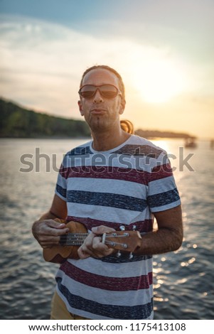 Happy young musician has a great time at the beach. He is standing by the water, playing ukulele and singing. Sunset over water.