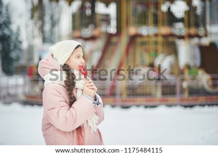 Girl in pink coat in snow Park. Girl plays in winter Park. Adorable child walking in snow winter forest. Snowy wheather. girl with lollipop rooster on background of old vintage carousel