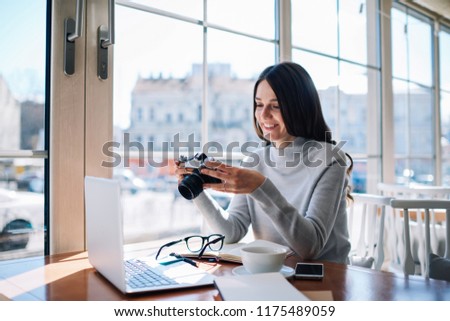 Positive female blogger photographer holding film camera in hands and checking settings sitting at coffee shop, successful cheerful amateur hipster girl happy from spending time during favourite hobby
