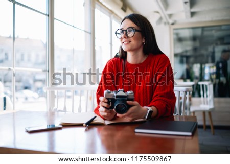 Good looking smiling hipster girl holding old fashioned technology while spending free time for enjoying hobby at cafeteria, positive female photographer in eyeglasses resting on leisure indoors