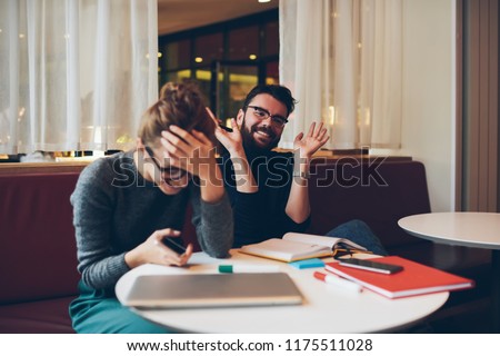 Cheerful young woman laughing together with best male friend during funny conversation, two positive students talking about funny story and relaxing on couch in break of studying in cozy cafeteria
